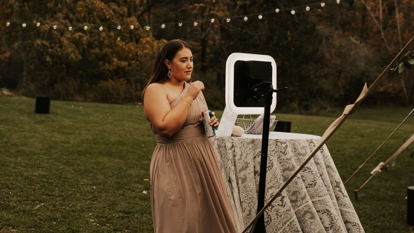 Une invitée en robe longue enregistre son message devant le vidéobooth, dans un jardin sous les guirlandes
